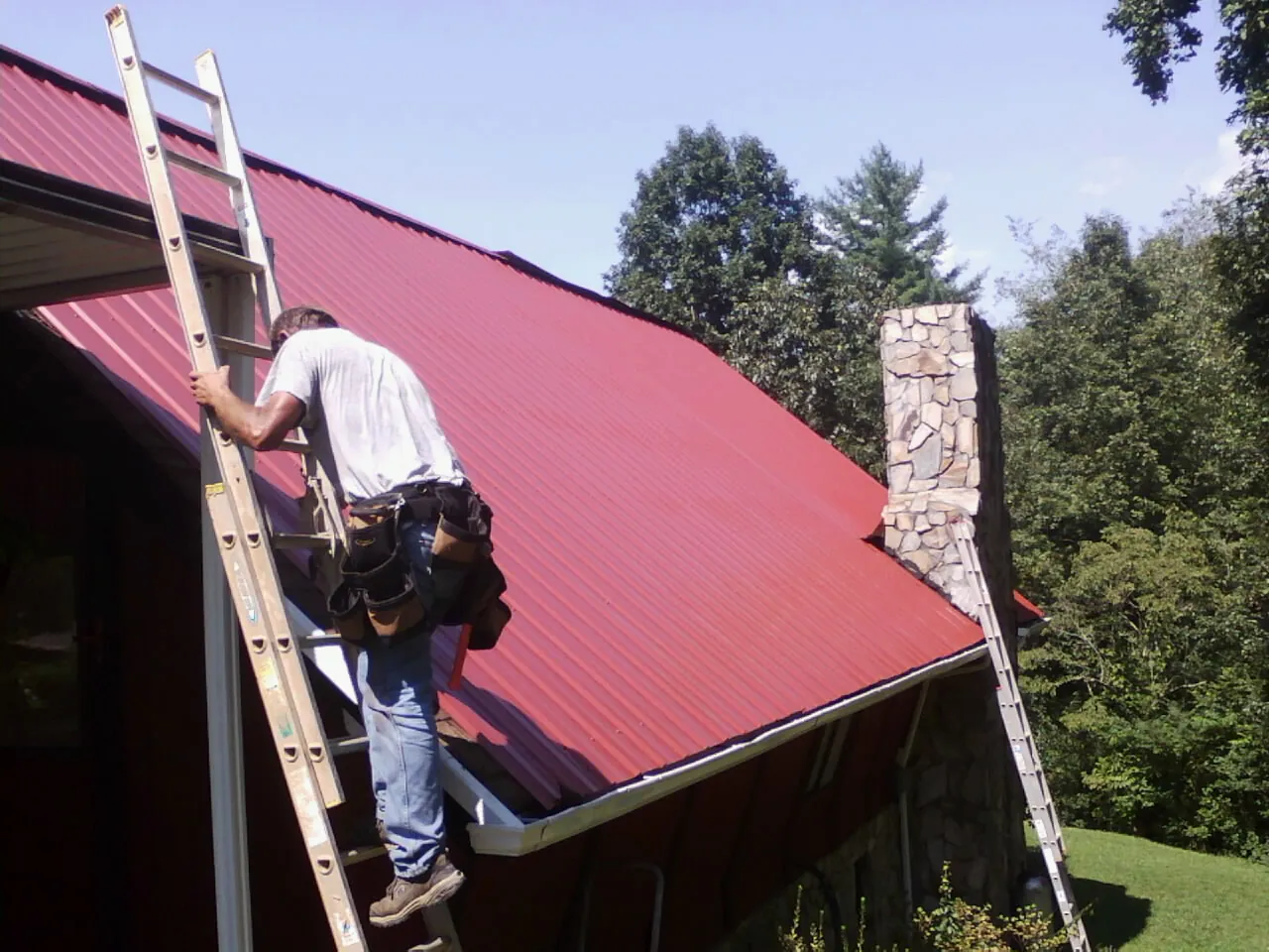 Roofer on a ladder beside a red metal roof and stone chimney — Soffit & Fascia Repair by Roofing Brothers in South Plainfield